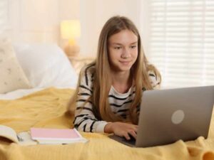 Estudiante en cama usando laptop y cuaderno, relacionado con desvelarse estudiando y falta de sueño.
